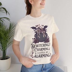 Peut inclure: Un t-shirt à manches courtes de couleur crème arborant un motif de chaudron sombre rempli de roses, de nœuds et d'ailes de chauve-souris. Sous le chaudron, le texte indique : BEWITCHINGLY CHARMING & A LITTLE BIT ALARMING, avec des dessins de clé, potion et sablier.