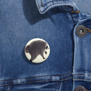Op de afbeelding: Een ronde pinback-button met een grafische illustratie van een donkergrijze wasbeer die loopt op een lichtcrèmekleurige achtergrond. De button is bevestigd aan een blauw spijkerjack met zichtbare stiksels en donkere metalen knopen.
