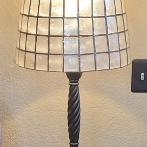 May include: A table lamp featuring a tapered drum shade constructed from square, translucent white shell tiles arranged in a grid pattern with dark grout lines. The lamp has a black, twisted metal stem and a square, tiered base with a power cord.