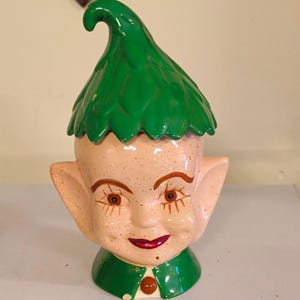 May include: A ceramic cookie jar shaped like an elf head with a speckled peach-colored face, large pointed ears, and a glossy green hat with a curled tip. The elf has painted brown eyes, long eyelashes, and red lips, wearing a green collar with a button.