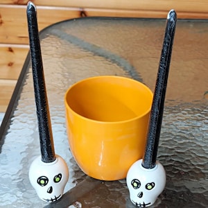 Puede incluir: Dos velas cónicas negras insertadas en portavelas blancos con forma de calavera y detalles oculares verdes, situadas a ambos lados de un cuenco naranja brillante, redondo y liso. Las velas tienen una superficie texturizada y puntas blancas.