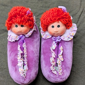 Könnte beinhalten: Ein Paar flauschige violette Hausschuhe mit Puppenköpfen und leuchtend rotem Wollhaar. Jeder Hausschuh hat einen gerüschten Kragen mit Blumenmuster, eine kleine violette Schleife am Hals und einen vertikalen Streifen aus passendem gerüschtem Stoff.