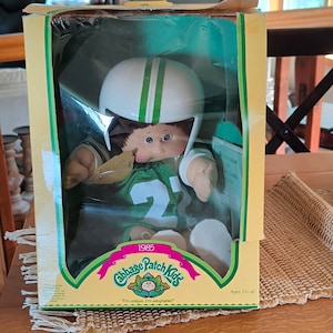 Puede incluir: Una muñeca Cabbage Patch Kids de 1985 en su embalaje original. La muñeca tiene cabello castaño, ojos azules y lleva un casco de fútbol americano blanco con una franja verde y una camiseta verde con el número 22. La caja muestra el logotipo.
