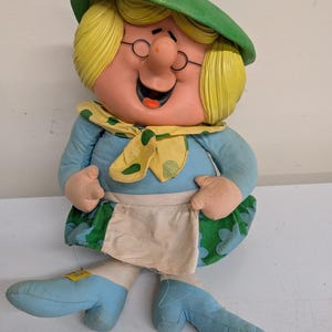 May include: A plush doll featuring a character with blonde hair, a green hat, and round glasses. The doll wears a light blue long-sleeved top, a yellow scarf with green polka dots, a green skirt with a floral pattern, and a white apron over light blue pants.