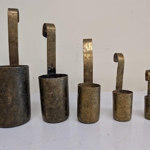 Op de afbeelding: Een set van vijf gegradueerde koperen maatbekers, elk met een cilindrisch lichaam en een lange, platte, gehaakte handgreep. De metalen oppervlakken vertonen een donkere, verouderde patina met zichtbare markeringen en tekst in de handgrepen.