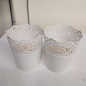 May include: Two white cylindrical plant pots featuring a decorative die-cut lace pattern along the upper rim. The pots have a smooth, solid base and a scalloped edge design at the top, providing a consistent look for both containers in this set.