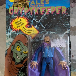 Peut inclure: Une figurine Tales from the Cryptkeeper dans son emballage d'origine. Le vampire porte un costume bleu et une cape violette. La carte présente un soleil jaune, une maison hantée et le texte Special Action Feature! Cape moves up Fangs come down.
