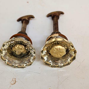 Può includere: Due pomelli per porta in vetro trasparente sfaccettato con una forma a stella a più lati. Ogni pomello presenta un inserto circolare color oro al centro ed è fissato a un perno metallico con un meccanismo di chiusura in metallo color bronzo.