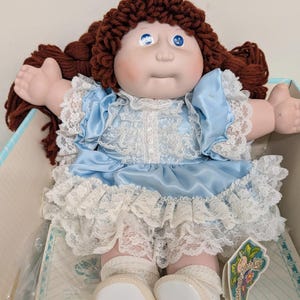 May include: A Cabbage Patch Kids doll with brown yarn hair, blue eyes, and a pale complexion. The doll wears a light blue satin dress with white lace trim on the sleeves and skirt, paired with white socks and white shoes, presented in a box.