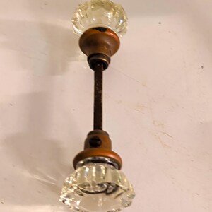 May include: A pair of clear, fluted glass door knobs connected by a central metal spindle. Each knob features a round, faceted glass head attached to a circular, dark bronze-toned metal base with a small set screw visible on the side of the neck.