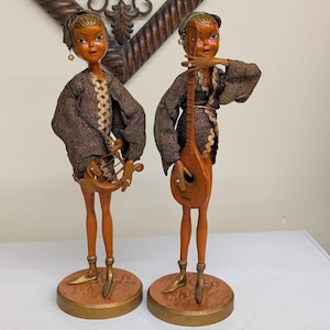 May include: Two standing figurines with orange skin tones, wearing dark textured jackets with gold trim and matching caps. One figurine holds a small wooden lyre, while the other holds a wooden lute. Both figures stand on round, gold-edged bases.