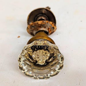 Könnte beinhalten: Ein Vintage-Türknauf mit einem klaren, facettierten Glasknauf und einem sternförmigen Muster in der Mitte. Der Knauf ist an einem angelaufenen Messingsockel und einer runden Metallmontageplatte befestigt, die sichtbare Anzeichen von Rost aufweist.