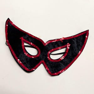 May include: A black velvet masquerade mask featuring a pointed, winged shape with vibrant red sequin trim outlining the outer edges and the eye openings. A thin black elastic strap is attached to the sides for securing the mask in place.