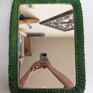 May include: A rectangular wall mirror featuring a thick, woven frame in a deep forest green color. The frame has a textured, braided appearance constructed from natural fibers, surrounding a clear, reflective glass surface with rounded corners.