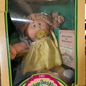 Puede incluir: Una muñeca Cabbage Patch Kids de 1985 con cabello de lana rubio en coletas, ojos verdes y un chupete amarillo. La muñeca viste un vestido amarillo y viene en una caja con un certificado de nacimiento y papeles de adopción, para mayores de 3 años.