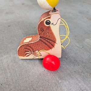 May include: A wooden pull toy shaped like a seal with a brown patterned body, a red wheel on the side, and a yellow and white ball balanced on its nose. The seal features a black eye, whiskers, and a thin yellow wire frame attached to its back.