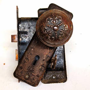 May include: A dark, weathered metal door knob and rectangular backplate set featuring intricate embossed floral patterns. The hardware includes a central keyhole, a round knob, and a heavy internal locking mechanism with visible rust and patina.
