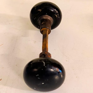 May include: A vintage door knob set featuring two round, glossy black knobs connected by a central square metal spindle. The knobs have a smooth, reflective surface with minor signs of wear, and the connecting rod shows a rusted, aged finish.
