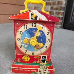 May include: A vintage Fisher-Price schoolhouse clock toy with a red frame, a yellow handle, and a central clock face featuring numbers 1 through 12. The dial displays a sun and moon graphic, and the base shows illustrations of children and a school.