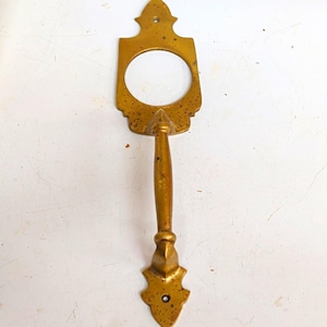May include: A brass door handle featuring a vertical, elongated design with a circular opening at the top and a pointed, decorative mounting plate at both the top and bottom. The metal surface shows a weathered, speckled patina throughout.