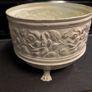 Puede incluir: Un cuenco de metal de color crema con un patrón en relieve de flores y hojas en el exterior. El cuenco tiene un acabado desgastado con reflejos dorados en los bordes y detalles, apoyado sobre tres pequeños pies ornamentados en garra.