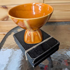 May include: A glossy orange ceramic bowl with a pedestal base and vertical yellow stripes, resting on a stack of three rectangular black coasters tied together with a thin piece of twine. The bowl has a wide, open rim and a smooth, reflective finish.