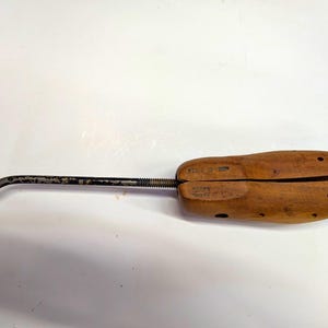 May include: A wooden shoe tree featuring a split-toe design with multiple ventilation holes and a metal heel rod. The rod includes a threaded section and a curved handle at the end. The wood has a natural brown finish with visible grain patterns.