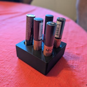 Może przedstawiać: Czarny kwadratowy stojak mieszczący cztery cylindryczne tubki kremu do ust. Tubki mają czarne nakrętki i etykiety z tekstem, w tym NYX Soft Matte Lip Cream. Pojemniki są ustawione pionowo w otworach matowej czarnej podstawy.
