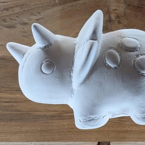 May include: A white, unpainted ceramic triceratops figurine featuring a smooth surface with molded stitch-like details along the edges of the head frill, body, and legs, and circular embossed patterns on the back and side of the dinosaur.