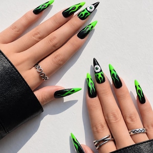 May include: Long, stiletto-shaped press-on nails featuring a black base with vibrant neon green flame patterns. Two of the nails include a detailed white and green eyeball graphic. The set is displayed on fingers adorned with silver rings.