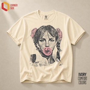 Peut inclure: Un t-shirt à manches courtes ivoire avec une impression graphique en noir et blanc représentant une personne aux cheveux tressés avec des pompons roses, soufflant une bulle de chewing-gum rose. Col rond et étiquette de taille M au col.
