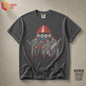 Può includere: Una t-shirt a maniche corte color grigio antracite con una stampa grafica della band Devo. Il design mostra cinque figure con abiti coordinati e cappelli rossi, con la parola DEVO in lettere bianche sotto una cupola rossa. Girocollo.