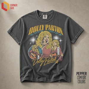 Peut inclure: Un t-shirt gris anthracite à manches courtes avec une impression graphique de Dolly Parton. Le motif comprend le texte "DOLLY PARTON" en jaune, des illustrations de la chanteuse avec une guitare et un micro, et des étoiles sur le devant.