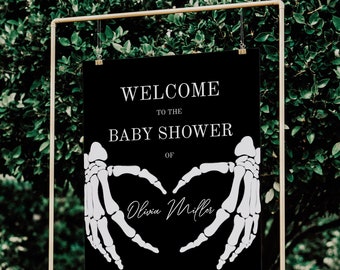 May include: A black baby shower welcome sign with white text and skeleton hand graphics. The sign reads "WELCOME TO THE BABY SHOWER OF Olivia Miller" and includes the location and date.