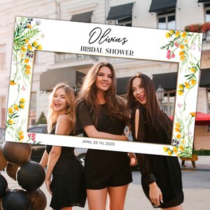 May include: A white photo booth frame with a gold border and floral accents. The frame has the text "Olivia's Bridal Shower" and "April 25th, 2029" printed on it.