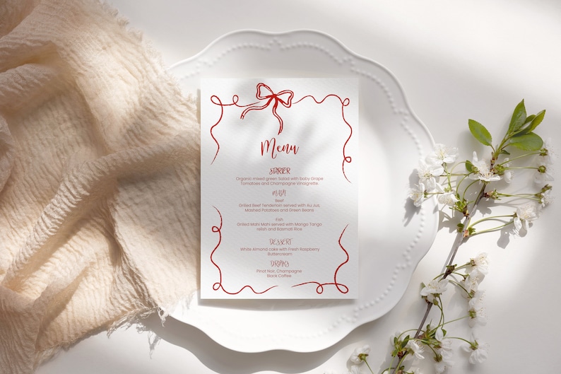 May include: A white menu card with a red ribbon design. The menu includes a starter, main course, dessert, and drinks. The text on the menu reads: "Organic mixed green salad with baby grape tomatoes and champagne vinaigrette", "Grilled Beef Tenderloin served with Au Jus, Mashed Potatoes and green beans", "Grilled Salmon served with Mango Tango salsa and Rosemary Rice", "White almond cake with fresh raspberry buttercream", "Red Wine, Champagne, Black Coffee".