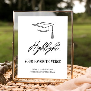 May include: A gold-framed sign with a white background and black text. The sign says "Highlight Your Favorite Verse" and includes a graduation cap illustration. It also says "Leave a post-it note of encouragement for Olivia."