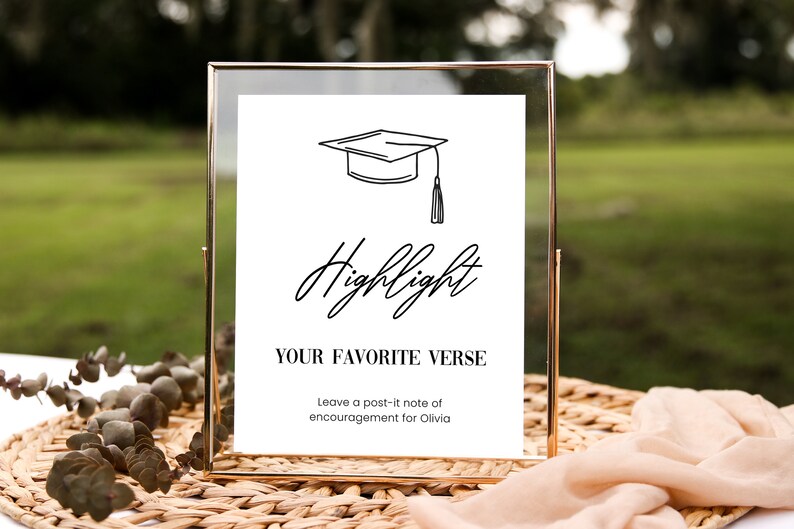 May include: A gold-framed sign with a white background and black text. The sign says "Highlight Your Favorite Verse" and includes a graduation cap illustration. It also says "Leave a post-it note of encouragement for Olivia."