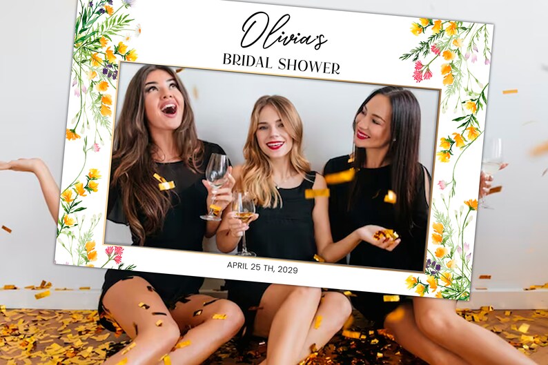 May include: A white photo booth frame with a floral border and gold trim. The text "Olivia's Bridal Shower" and "April 25th, 2029" is printed on the frame. Three women are posing inside the frame.