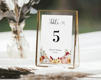May include: A gold-framed table number card with a white background and black text. The card reads "Table 5" and "Olivia & Thomas 10.22.2023". The bottom of the card features a watercolor illustration of fall-themed flowers and pumpkins.