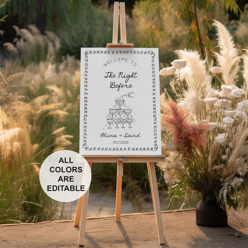 May include: A white sign with black text and a graphic of champagne glasses. The sign reads "Welcome to The Night Before Olivia + David 05.21.2035". The sign is on a wooden easel in front of a grassy background.