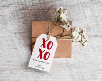 May include: A white gift tag with red "XO" lettering and the text "Happy Valentine's Day, Love, The Millers" tied to a brown gift box with a sprig of white flowers.