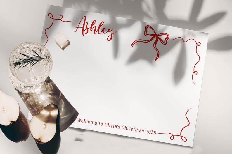 May include: A white card with red script that reads "Ashley" and "Welcome to Olivia's Christmas 2035." A glass of water with ice and a sprig of rosemary sits next to a sliced pear.