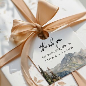 May include: A white thank you tag with a watercolor mountain scene and the text "thank you" in black cursive. The tag also includes the text "For celebrating with us FIONA + JASON".
