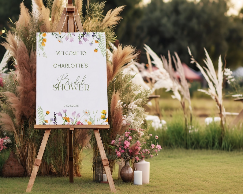 May include: A white sign with a floral border and the text "Welcome to Charlotte's Bridal Shower 04.25.2025" displayed on an easel in a grassy outdoor setting.