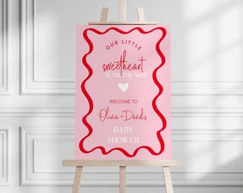 May include: Pink baby shower sign with a red wavy border. The sign says "Our little sweetheart is on the way" and "Welcome to Olivia + David's Baby Shower".