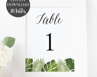 May include: White frame with a white card inside. The card has black text that says "Table 1" and a green border of tropical leaves.