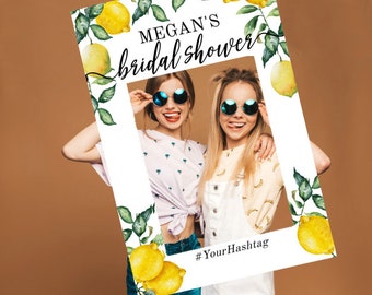 Può includere: Un accessorio per foto booth bianco a tema limone. L'accessorio ha un ritaglio per due volti e dice "MEGAN'S bridal shower" in alto. La parte inferiore dell'accessorio dice "#YourHashtag".