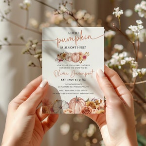 May include: A baby shower invitation with a fall theme. The invitation features a watercolor illustration of pumpkins and flowers in shades of orange, pink, and brown. The text reads "A little pumpkin is almost here!" and includes the date, time, and location of the shower. The invitation is perfect for a fall baby shower.
