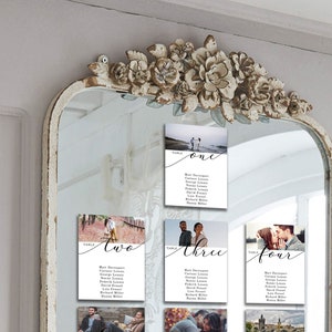 May include: A white mirror with a decorative floral frame, displaying four white cards with photos and seating arrangements for a wedding reception. The cards are labeled "Table One", "Table Two", "Table Three", and "Table Four".
